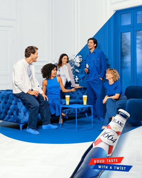 Group of five adults socializing in blue-themed room with beer; Blanc 1664 bottle in the foreground with white and blue brand design.