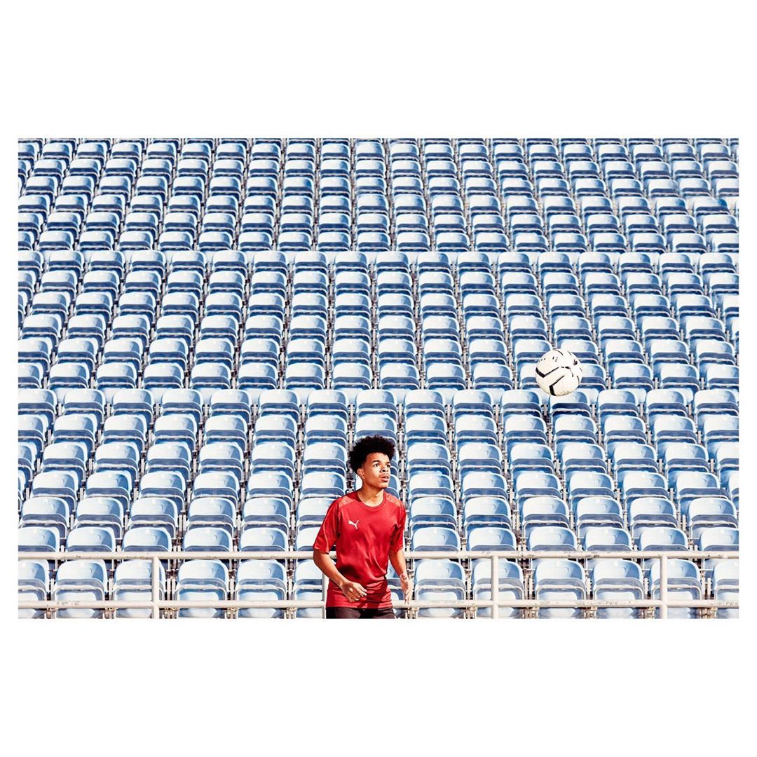 Teenage boy in a red shirt practices soccer in an outdoor stadium with rows of blue seats and a soccer ball floating mid-air.