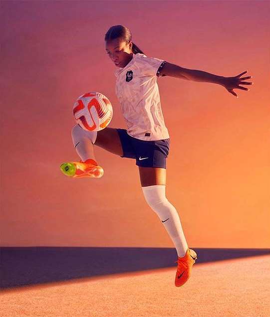 A female soccer player wearing France national uniform balances a soccer ball on her foot, mid-air with orange background.