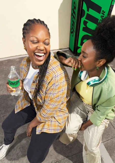 Two young Black women laugh together while sitting. One holds a Sprite bottle near a Sprite cooler in an urban setting.