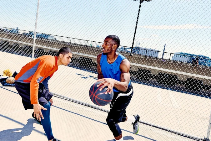 Two adult males play basketball outdoors on an urban court; one dribbles a ball while the other faces him in a defensive stance.