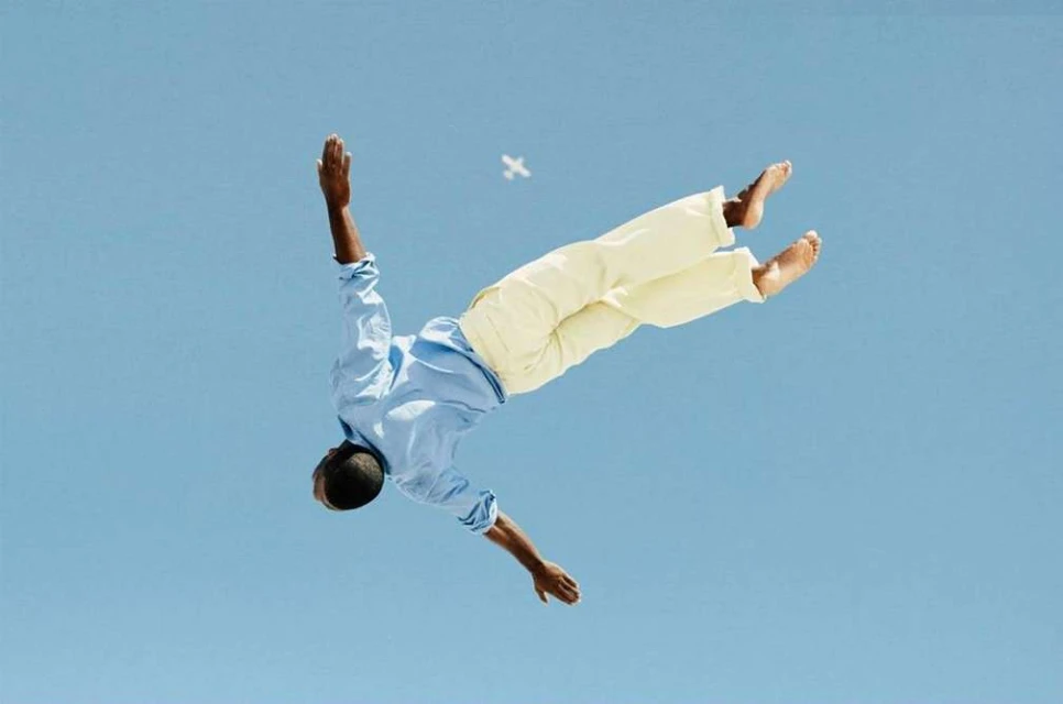 Man in blue shirt and yellow pants appears to float mid-air in front of a clear blue sky with arms and legs outstretched.