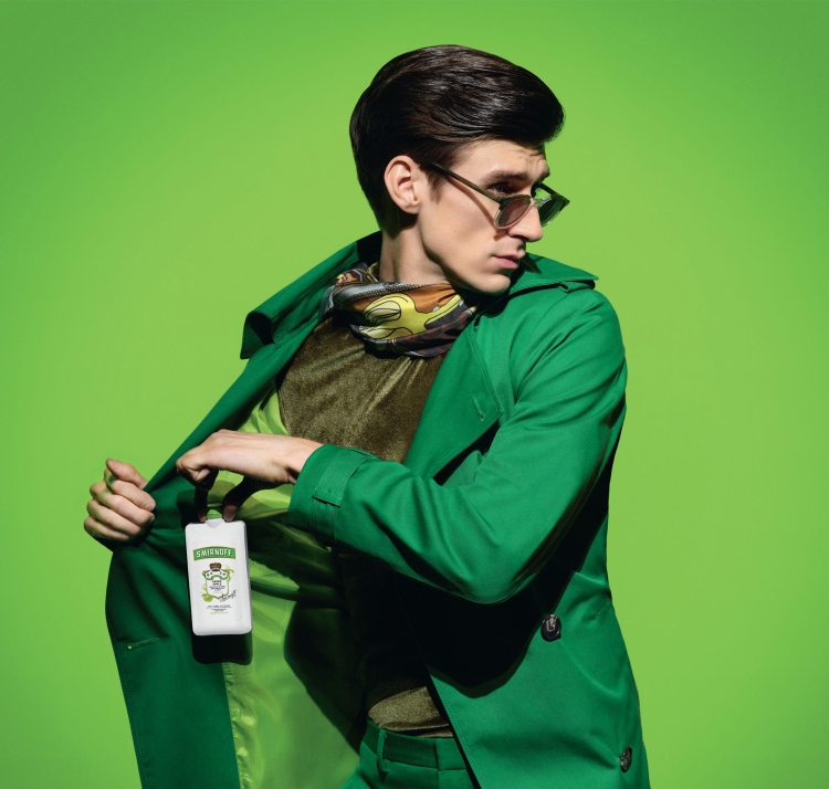 An adult male in green coat and glasses holds a white bottle against a bright green backdrop, looking to his left in a studio shot.