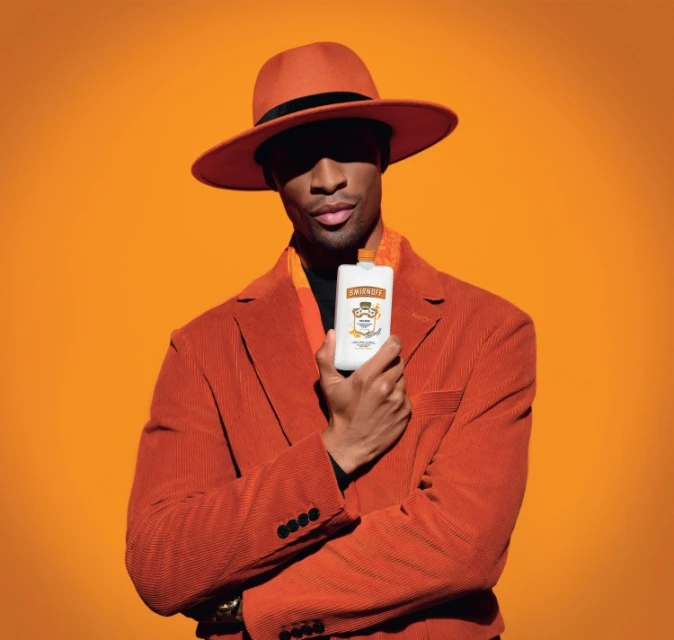 Stylish African-American man in hat and corduroy suit holds Smirnoff Ice pouch against an orange background in a portrait shot.