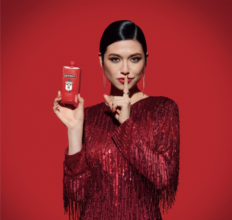 Adult woman in red dress holds a Smirnoff bottle up with a finger to her lips against a matching red background in studio lighting.