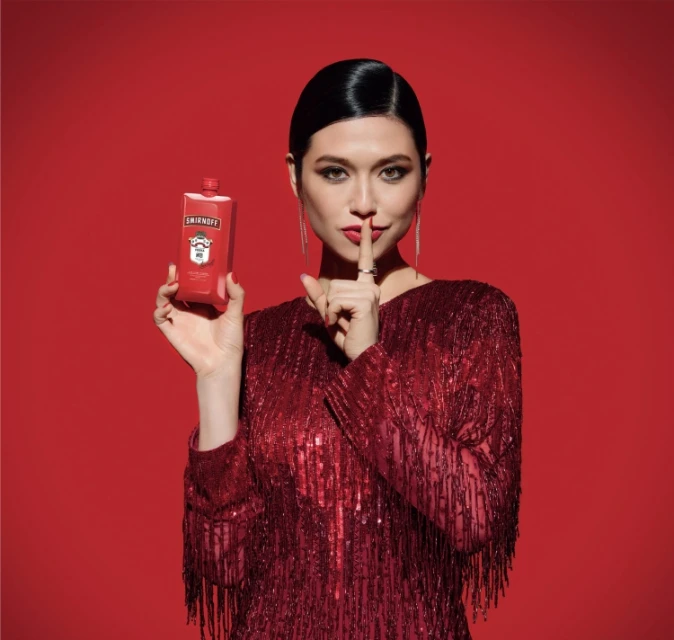 Adult woman in red dress holds a Smirnoff bottle up with a finger to her lips against a matching red background in studio lighting.
