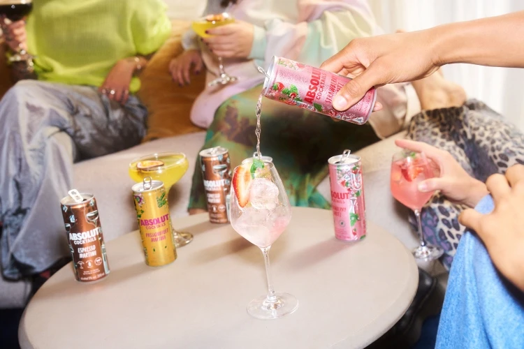 Person pours Absolut cocktail into a stemmed glass with ice and strawberries, surrounded by various Absolut cocktail cans and other people.