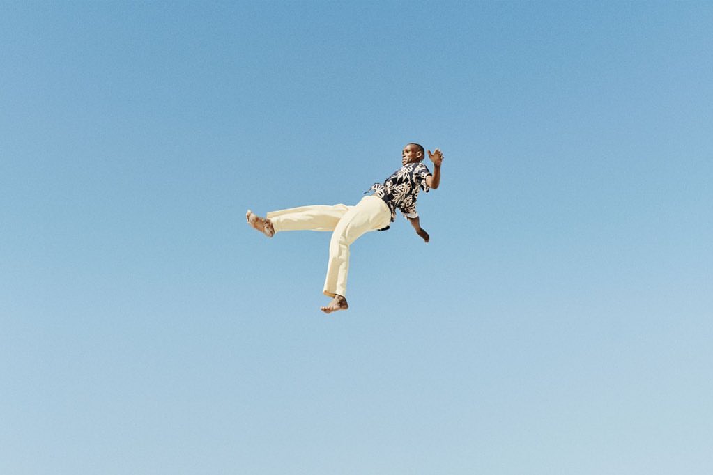 A Black adult male in mid-air wearing a patterned shirt and tan pants against a clear blue sky backdrop in a dynamic pose.