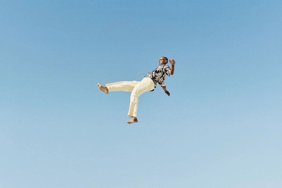 A Black adult male in mid-air wearing a patterned shirt and tan pants against a clear blue sky backdrop in a dynamic pose.