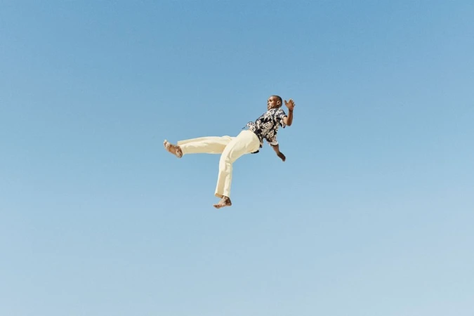 A Black adult male in mid-air wearing a patterned shirt and tan pants against a clear blue sky backdrop in a dynamic pose.