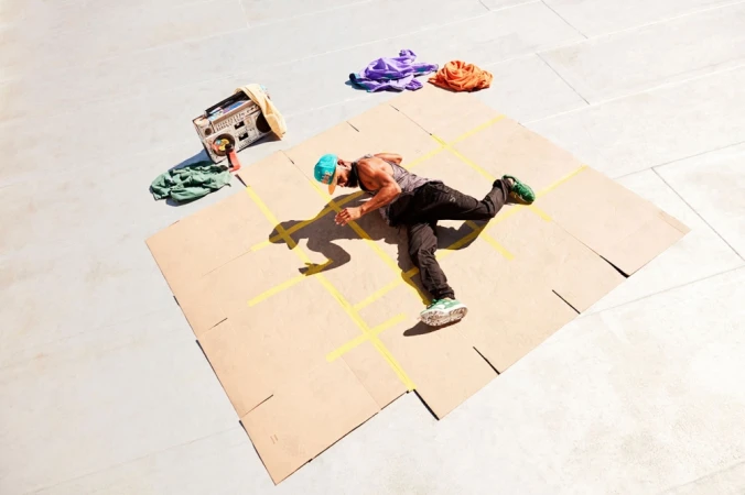 Male dancer performs a routine on a taped cardboard square outdoors, wearing a hat, tank top, and athletic pants, with a boombox nearby.