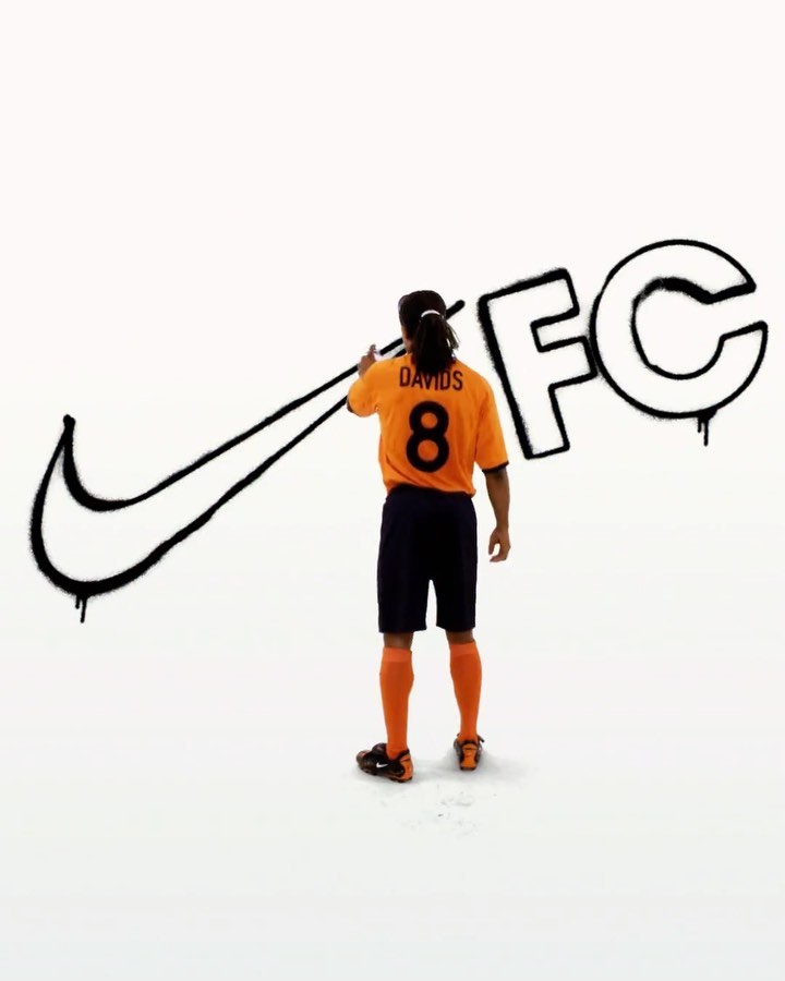 Soccer player in orange jersey spray paints FC and the Nike swoosh on white wall, promoting sports brand and team spirit.