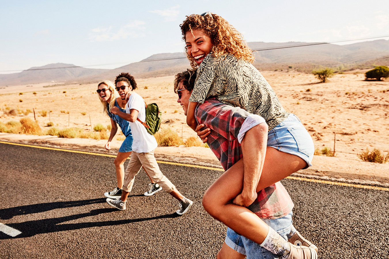 Four young adults walk along a rural road, with one woman riding piggyback, enjoying travel, friendship, and outdoor recreation.
