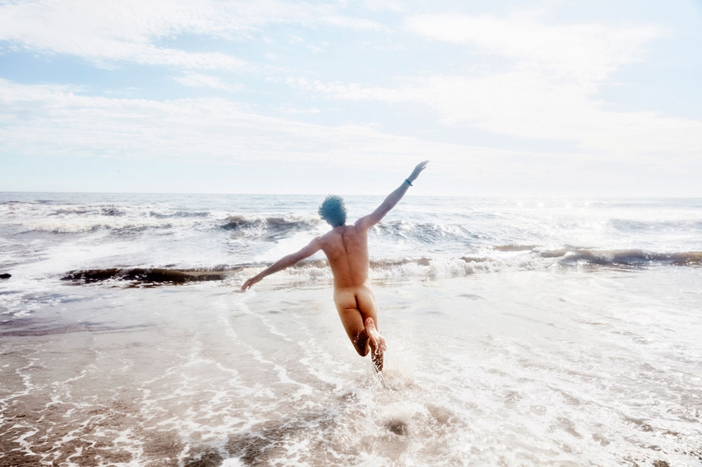 Back view of a nude adult jumping with arms outstretched at the beach on a sunny day with waves in the background.