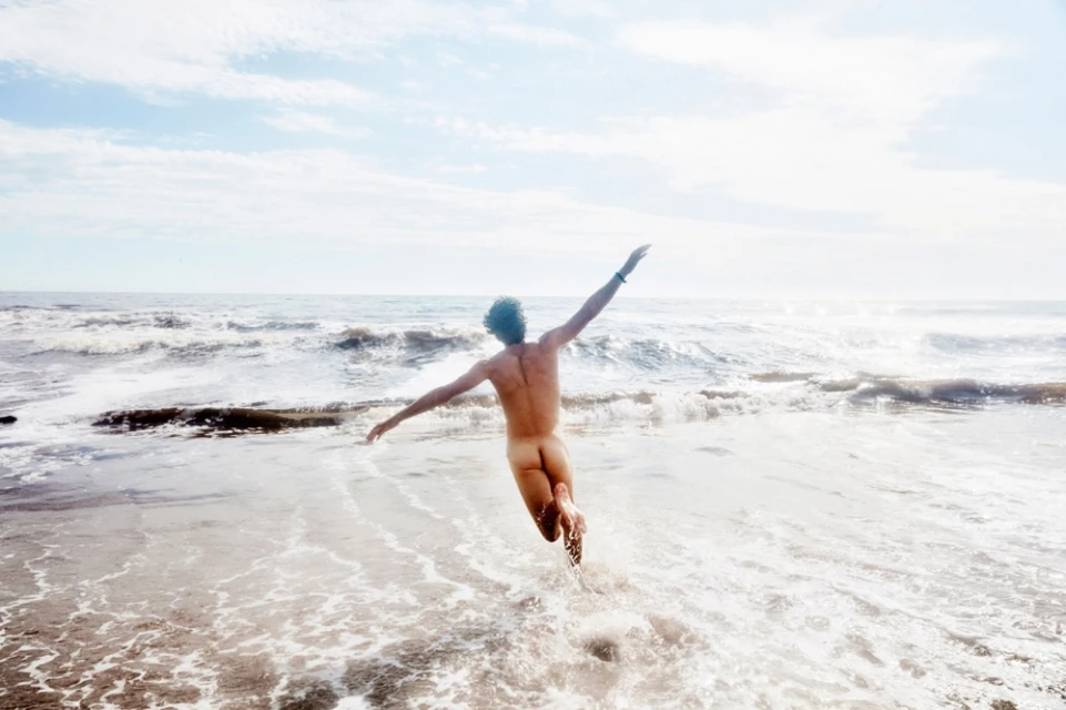 Back view of a nude adult jumping with arms outstretched at the beach on a sunny day with waves in the background.