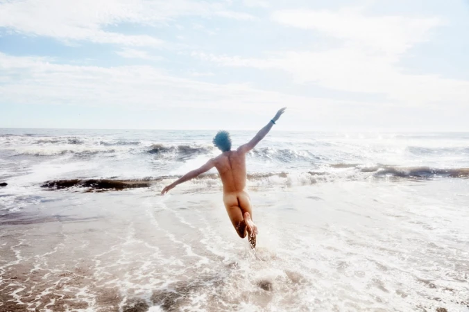 Back view of a nude adult jumping with arms outstretched at the beach on a sunny day with waves in the background.