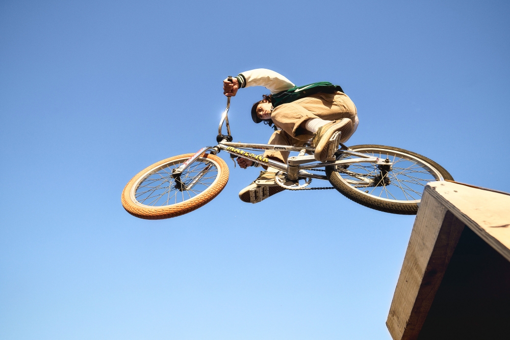 BMX rider mid-air performing a stunt on a bike against a blue sky, with a wooden ramp visible in the lower right corner of the frame.