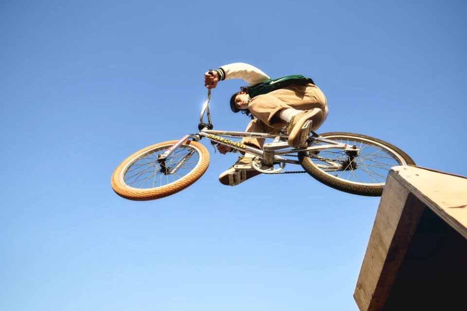 BMX rider mid-air performing a stunt on a bike against a blue sky, with a wooden ramp visible in the lower right corner of the frame.