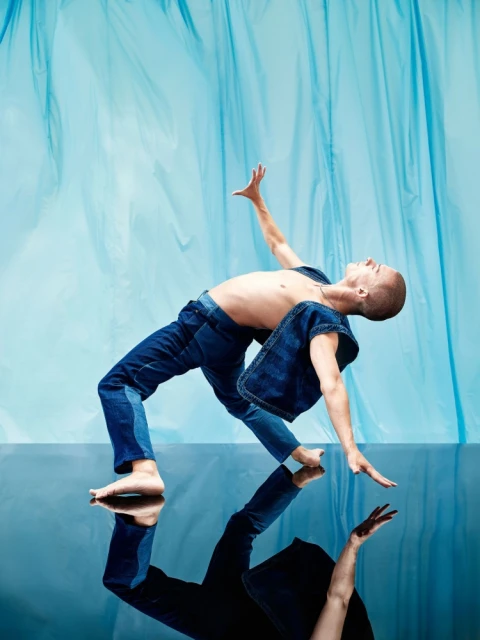 Male dancer in denim vest and jeans, strikes a dynamic pose on reflective surface against a draped blue background, captured in studio lighting.