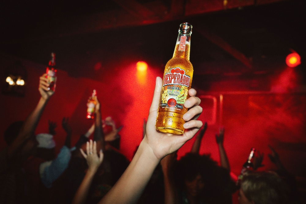 Close-up of hand holding Desperados beer at crowded indoor party with red lighting, celebrating with raised arms and beverage bottles.