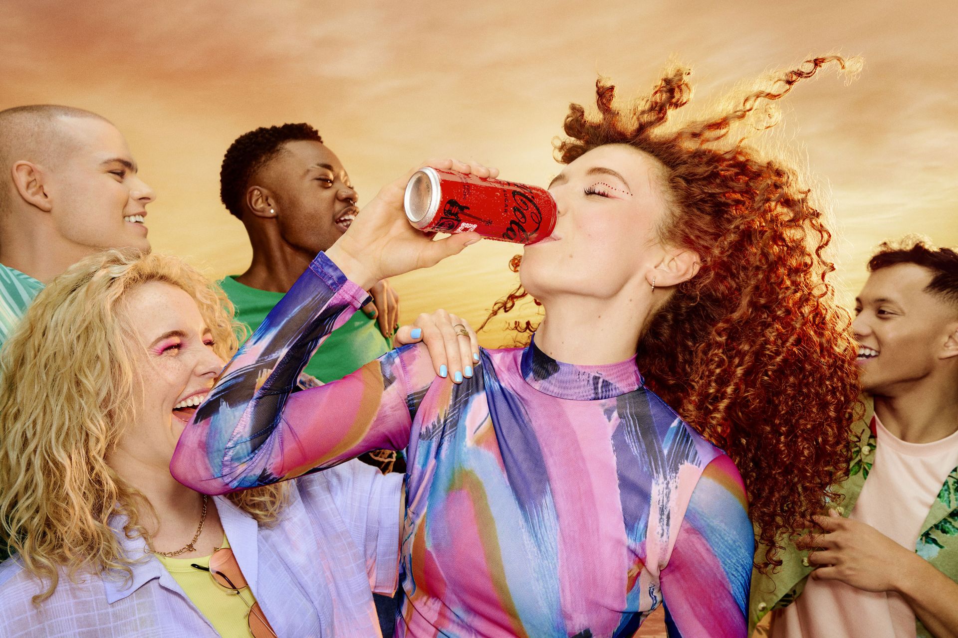 Group of six diverse young adults gathered outdoors, one woman drinking from a red can, wearing colorful clothing against a warm background.