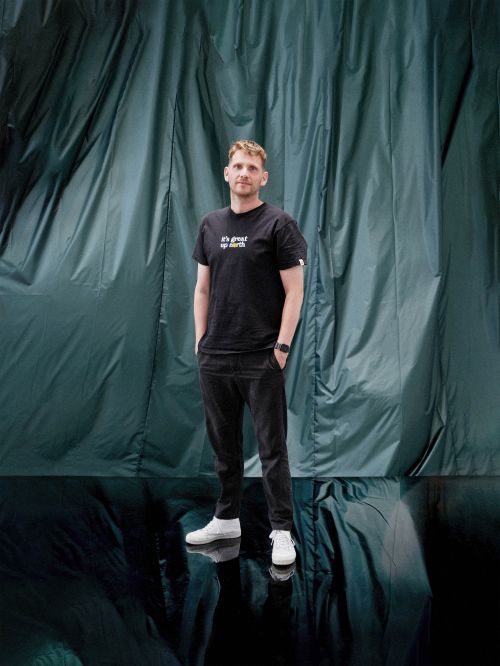 Full-length portrait of a man in a black T-shirt and black pants with hands in pockets standing against a draped blue-green fabric backdrop.