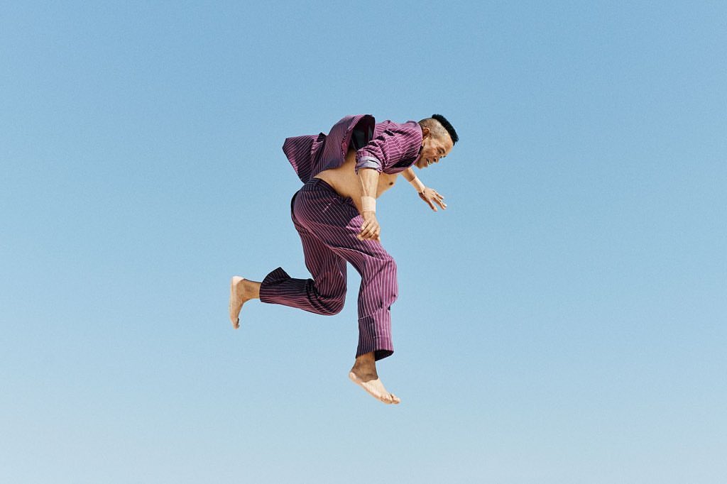 An adult male in a purple suit jumps against a light blue sky background, capturing a dynamic and unusual fashion-forward pose outdoors.