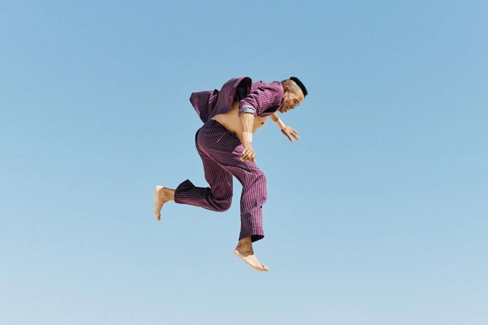 An adult male in a purple suit jumps against a light blue sky background, capturing a dynamic and unusual fashion-forward pose outdoors.