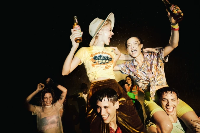 Enthusiastic group of young adults celebrate outdoors at night, illuminated by overhead light, holding beer bottles, smiling, and laughing.