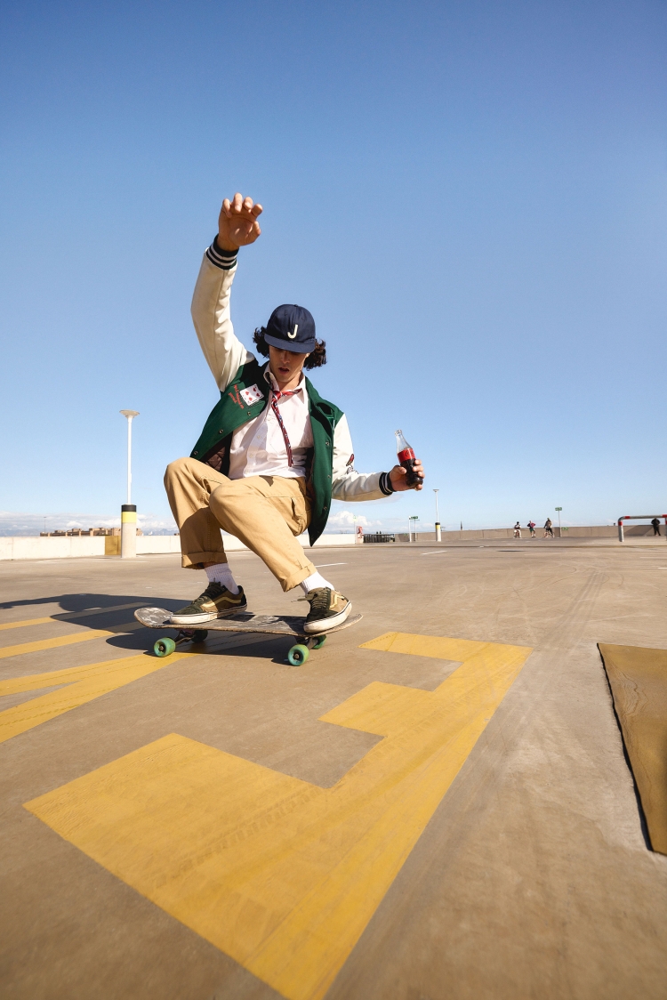 Young man skateboarding on a concrete surface outdoors under a clear blue sky, wearing a letterman jacket and holding a bottle.