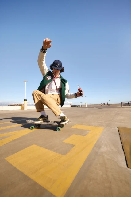Young man skateboarding on a concrete surface outdoors under a clear blue sky, wearing a letterman jacket and holding a bottle.