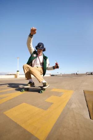 Young man skateboarding on a concrete surface outdoors under a clear blue sky, wearing a letterman jacket and holding a bottle.