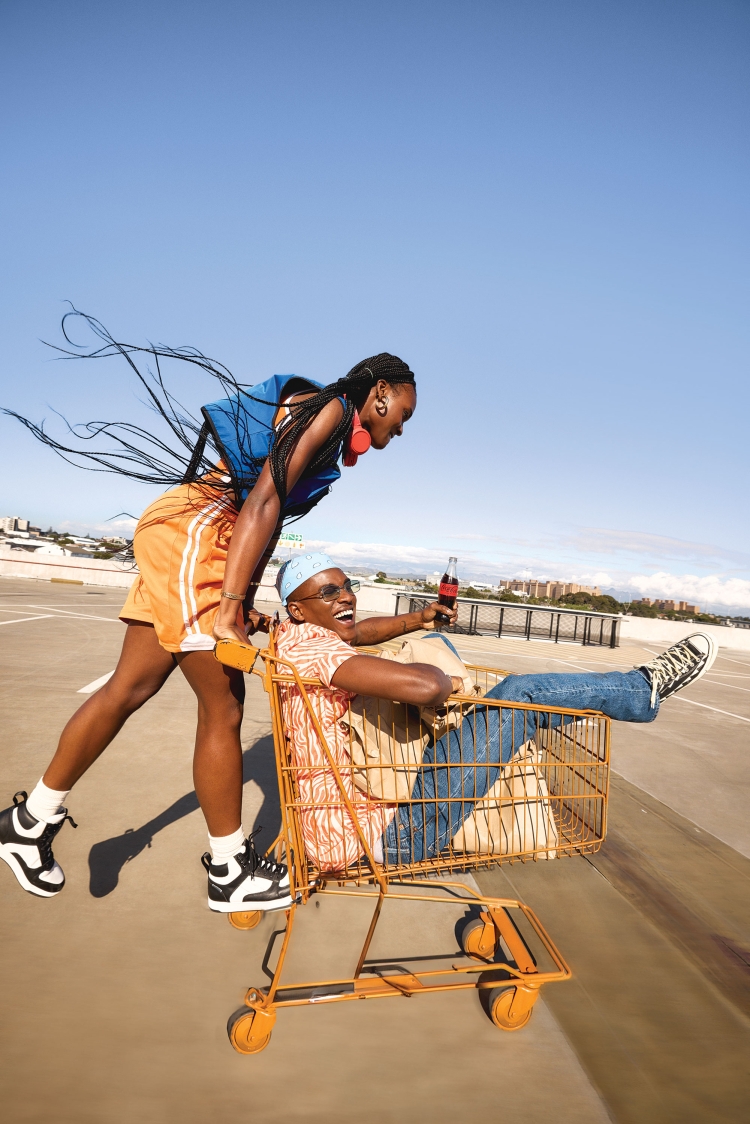 Two adults, one male and one female, on an outdoor parking deck. The female is pushing the male in a yellow shopping cart.