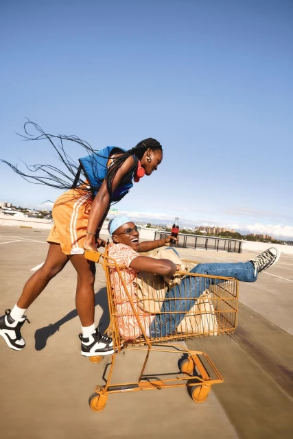 Two adults, one male and one female, on an outdoor parking deck. The female is pushing the male in a yellow shopping cart.