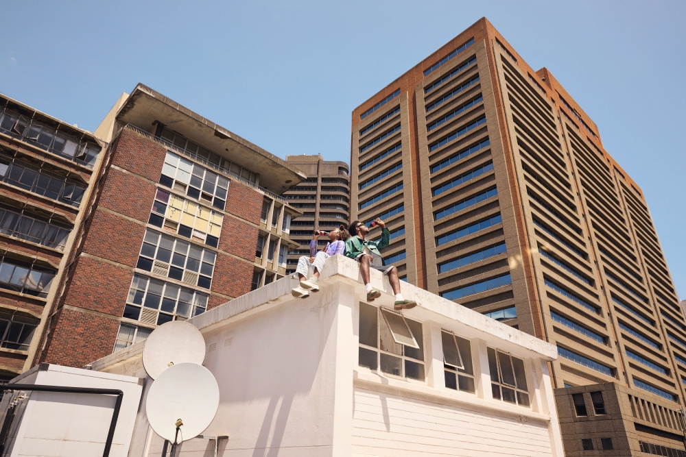 Two adults sit on a rooftop, drinking from bottles, with city buildings in the background under a clear sky, captured with a low-angle.