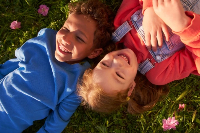 Two smiling children lying in the grass together among small pink flowers, viewed from overhead.