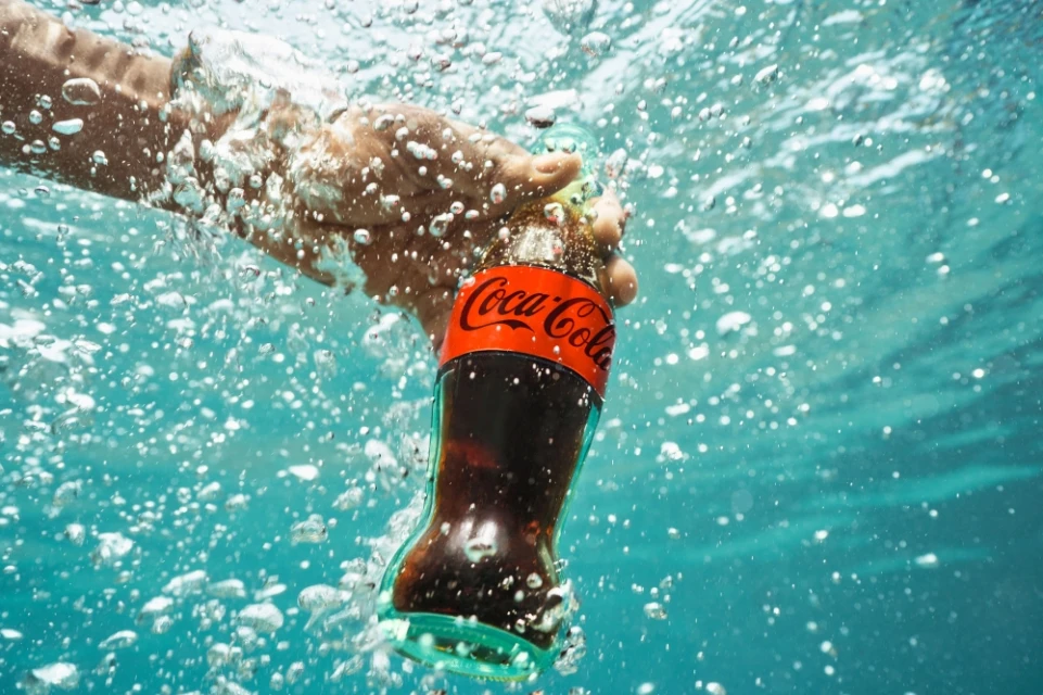 A hand holds a Coca-Cola bottle underwater amid bubbles, creating a refreshing visual with vibrant colors and dynamic composition.