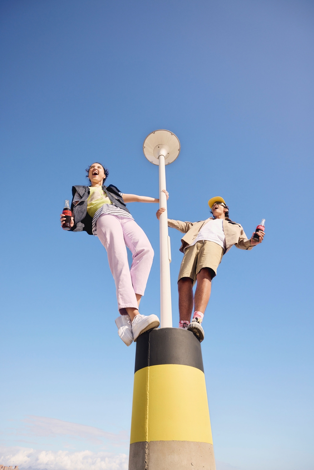 Two youthful figures playfully pose atop a marker, beverages in hand, against a bright blue sky backdrop, embodying freedom and joy.
