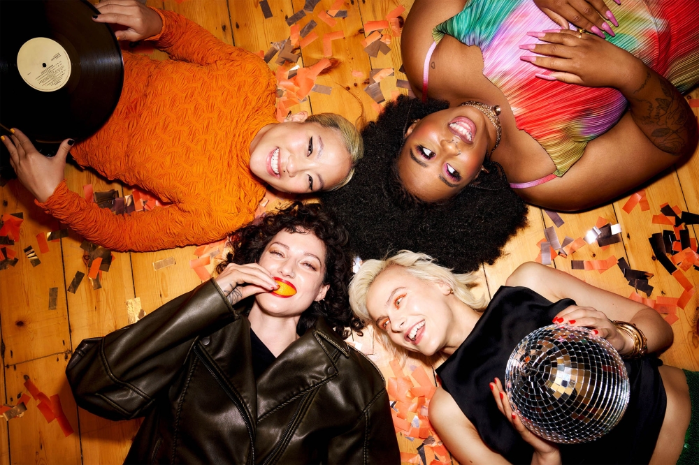 Four smiling young women laying on a wooden floor covered in confetti with a disco ball and vinyl record in a top-down shot.
