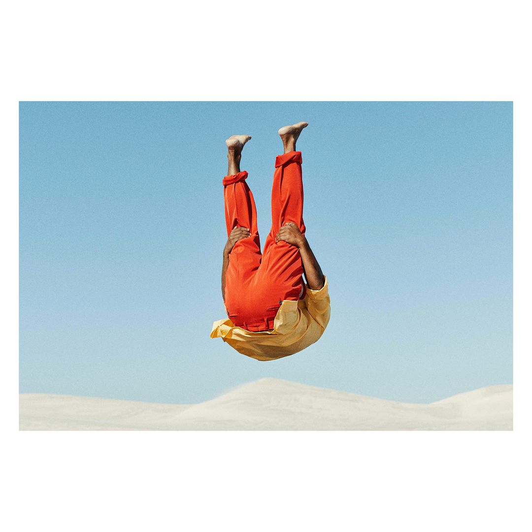 A person in mid-air doing a handstand in an outdoor setting with orange pants and a gold shirt against a clear blue sky.