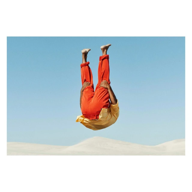 A person in mid-air doing a handstand in an outdoor setting with orange pants and a gold shirt against a clear blue sky.