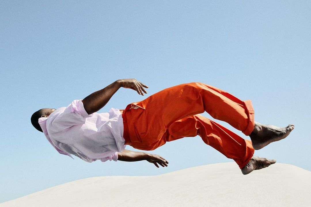 Adult man in bright orange trousers and light pink shirt levitates over a white sand dune against a clear blue sky in an outdoor setting.