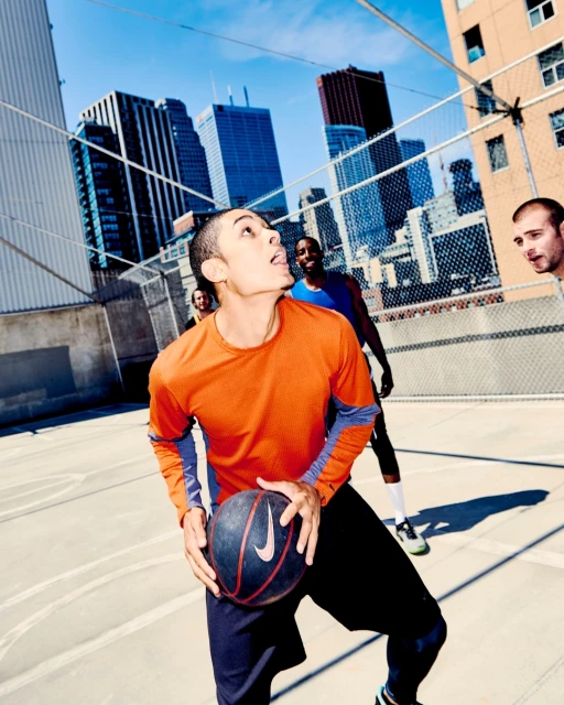 A low angle captures a young adult male basketball player in motion on an urban court, dribbling a basketball with buildings in the background.
