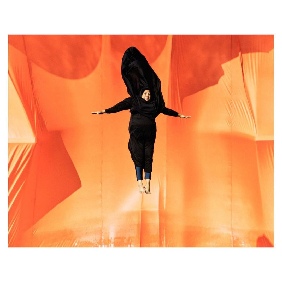 Person in black clothing jumps in front of an orange background. Arms are extended, creating a dynamic, full-body pose in mid-air.