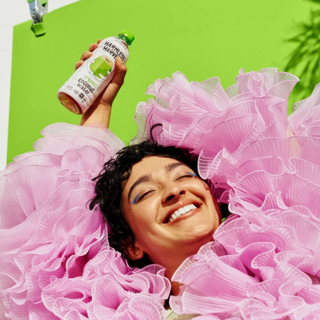 Woman in pink ruffles smiles, holding Harmless Harvest coconut water against a vibrant green background with tropical plant shadows.