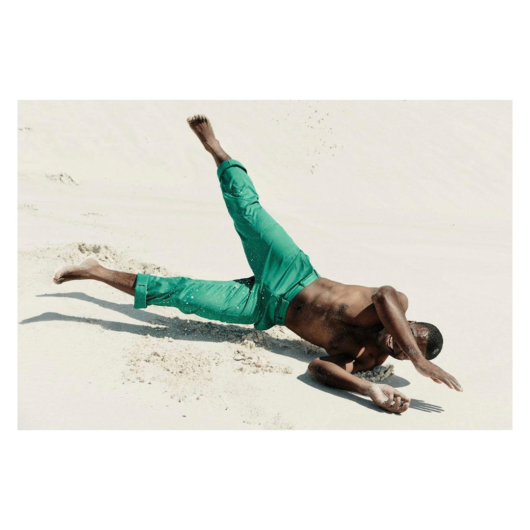 Adult male in a dynamic pose on a sandy beach wearing green pants, captured in a photograph with bright lighting and clear visibility.
