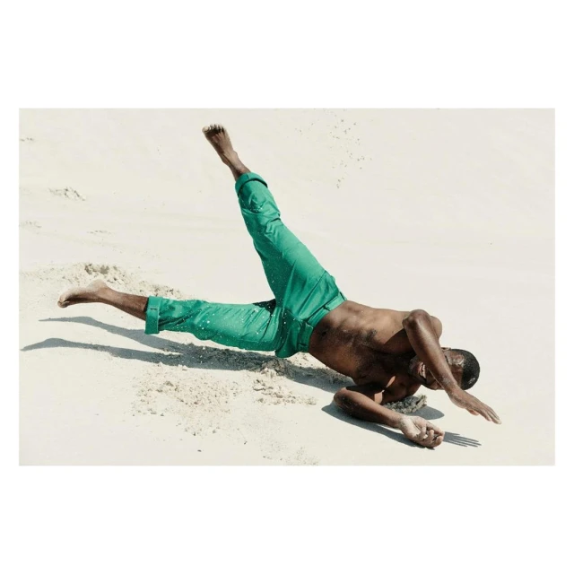 Adult male in a dynamic pose on a sandy beach wearing green pants, captured in a photograph with bright lighting and clear visibility.