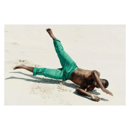 Adult male in a dynamic pose on a sandy beach wearing green pants, captured in a photograph with bright lighting and clear visibility.