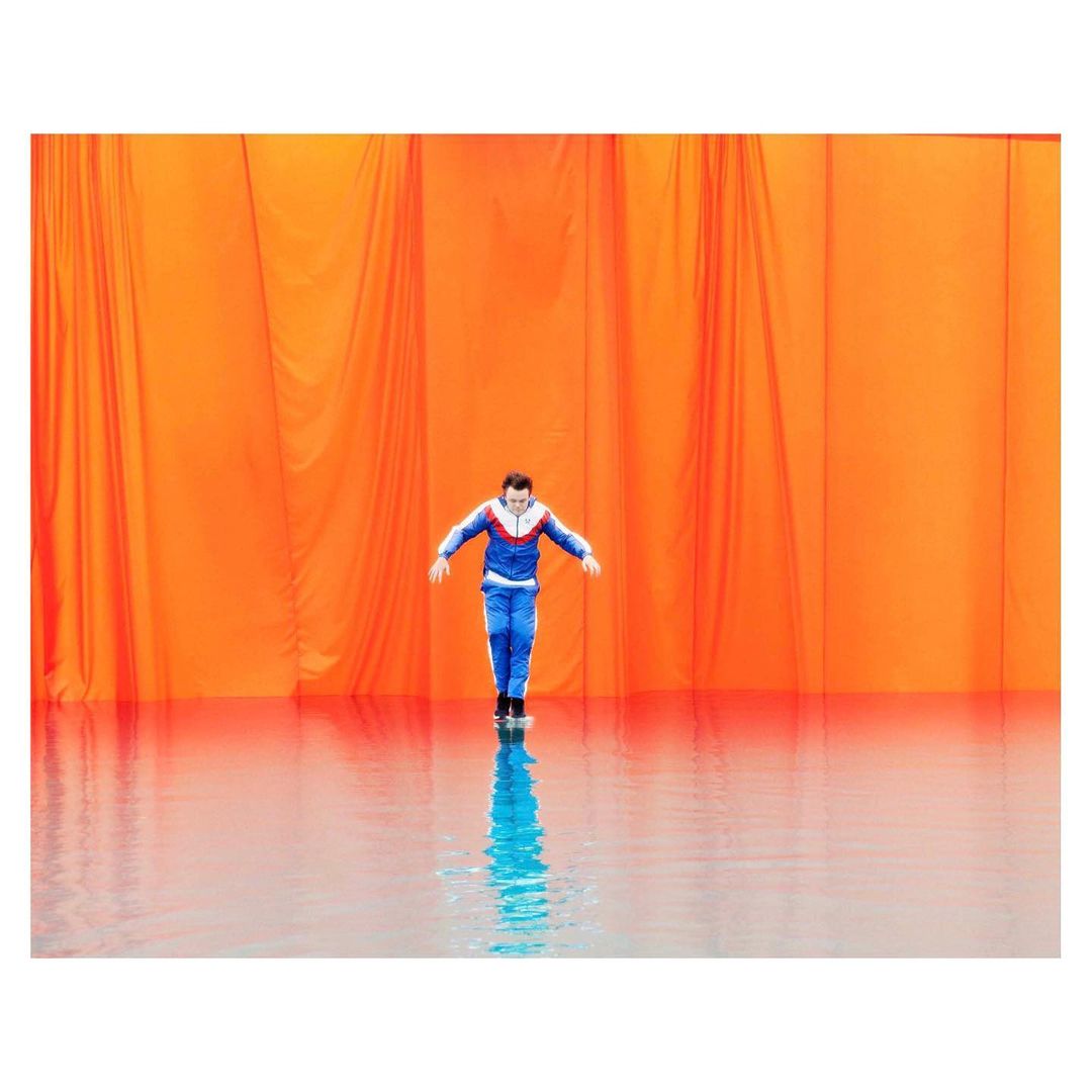 A man in a blue tracksuit stands on a wet surface in front of an orange curtain backdrop in a studio setting.