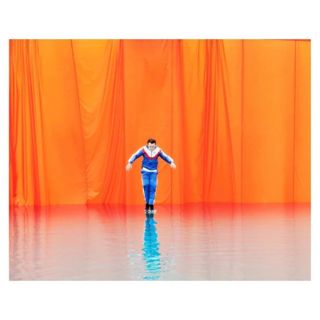 A man in a blue tracksuit stands on a wet surface in front of an orange curtain backdrop in a studio setting.